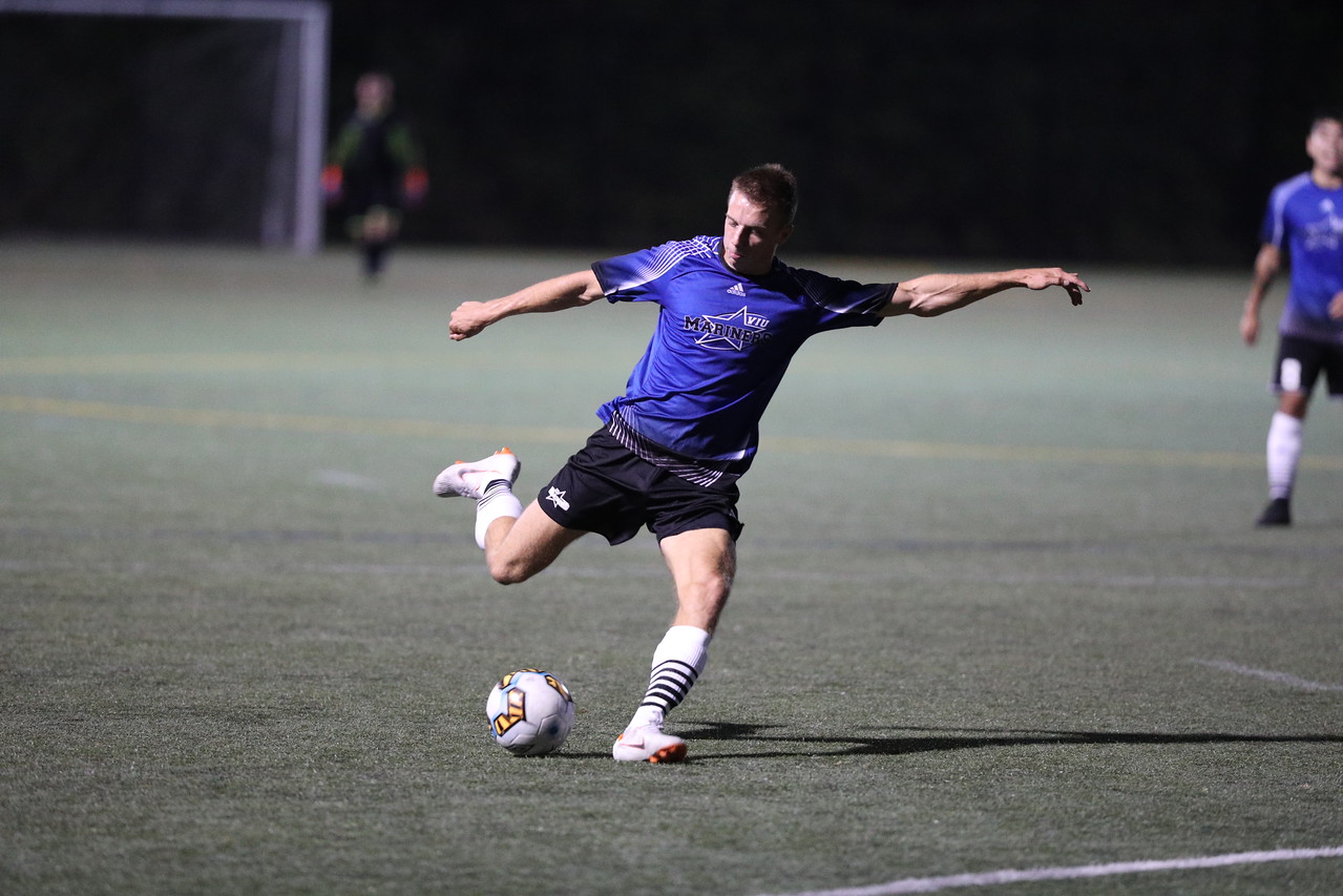 VIU men’s soccer team midfielder Griffin Douglas in mid-kick, playing in a game against Mid-Isle. He is center frame, wearing the team’s blue jersey and black shorts. Other players run in the background.
