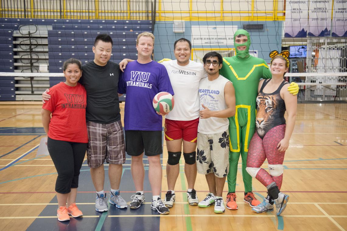 Seven members of one of the many intramural teams competing in a VIU volleyball tournament, standing shoulder-to-shoulder in front of a net in the VIU gym. One player is wearing a full green Kick-Ass costume, another is wearing a false nose and glasses mask, a third sports a tiger ears headband.