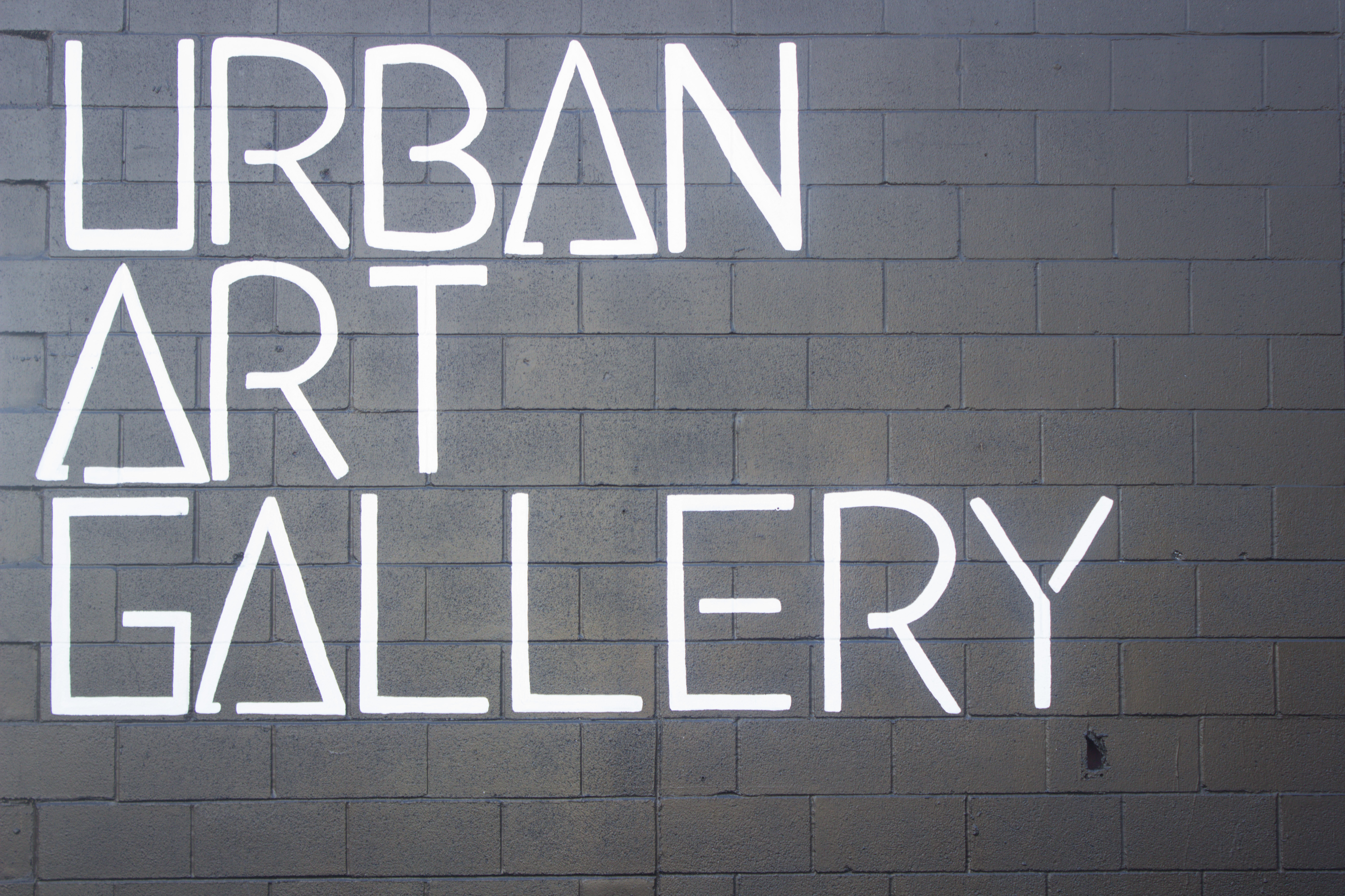Capitalized white letters spell out Urban Art Gallery, each word falls under the next so that first letters align. It’s written over a wall of cinder blocks painted black.