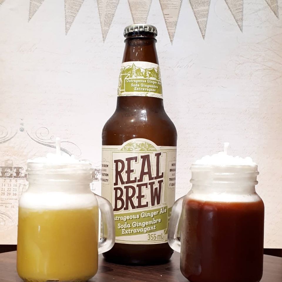A beer bottle with ‘REAL BREW’ written across in brown writing on the label sits in between a yellow and reddy-brown “beer mug” candle.