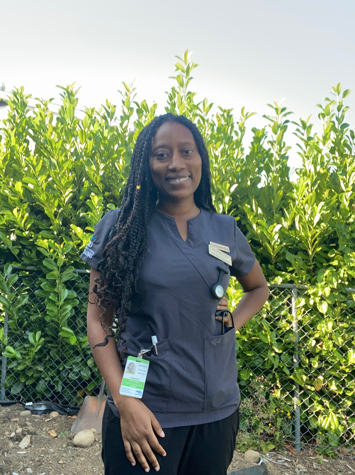 A nurse in grey scrubs smiles, standing in front of green bushes