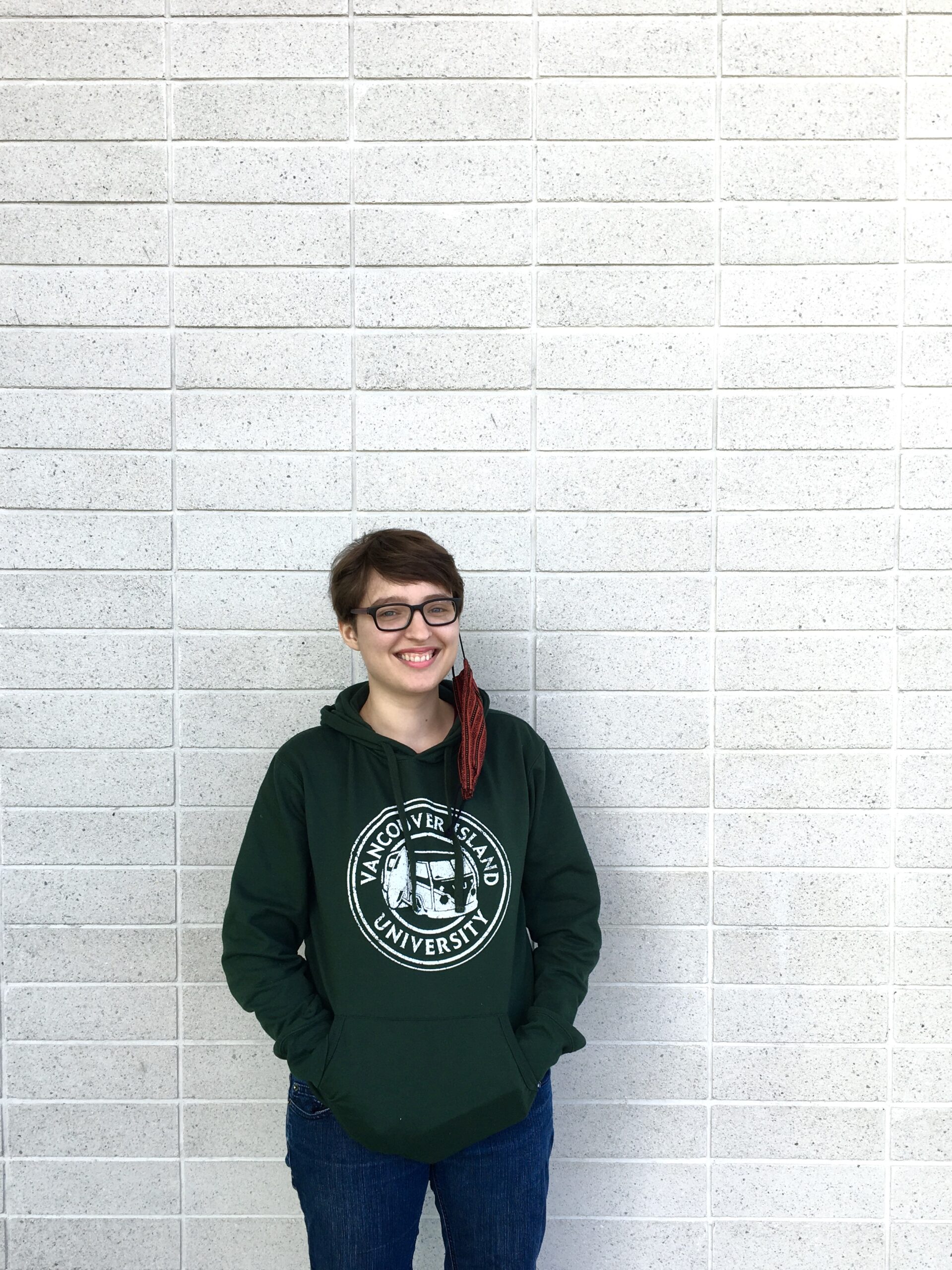 A woman in a green sweater smiles standing in front of a grey brick wall, she has a mask hanging from her left ear