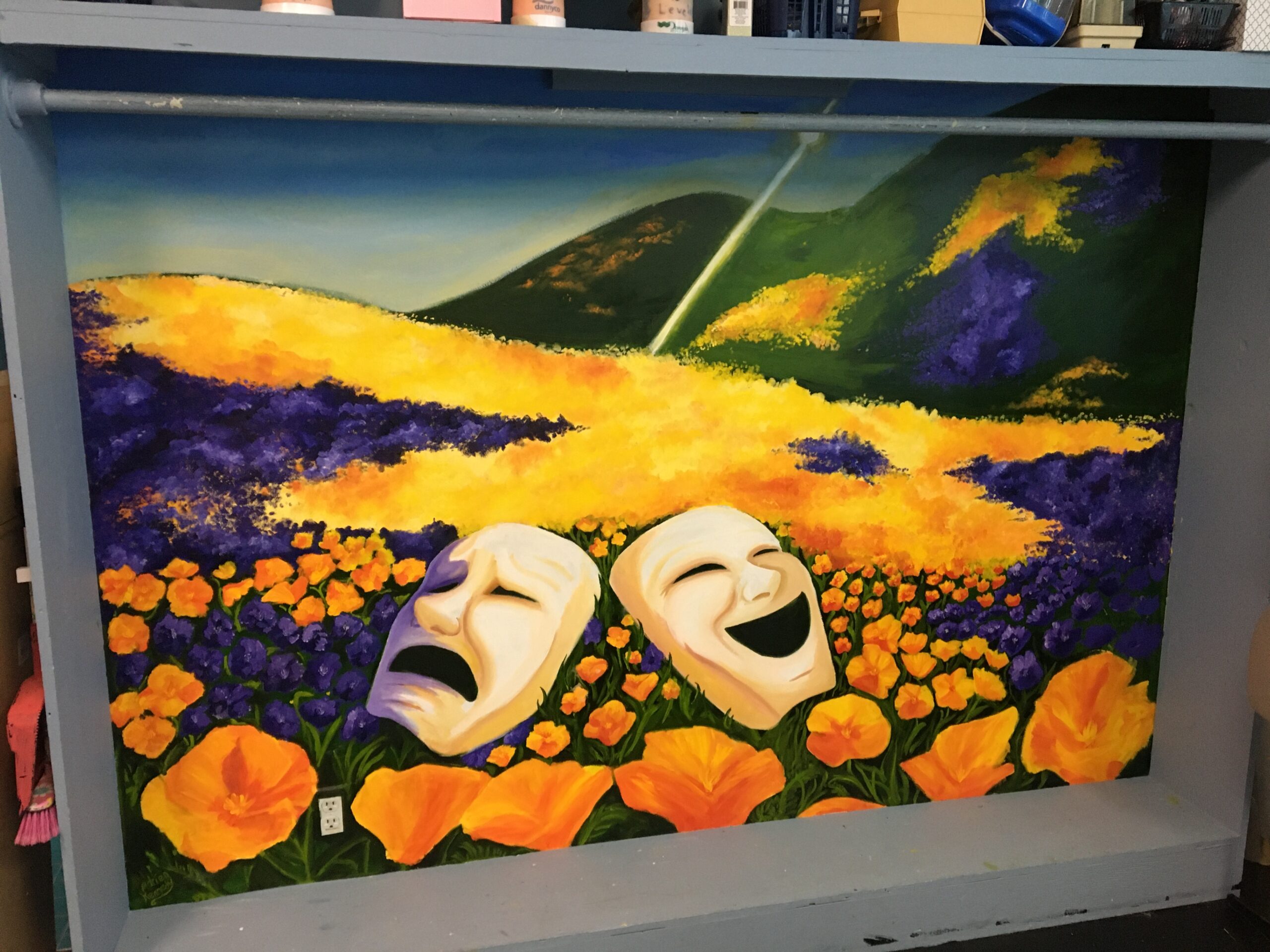 Student-painted mural in theatre dressing room: a comedic mask and a tragic mask lie in the forefront of a flowering meadow