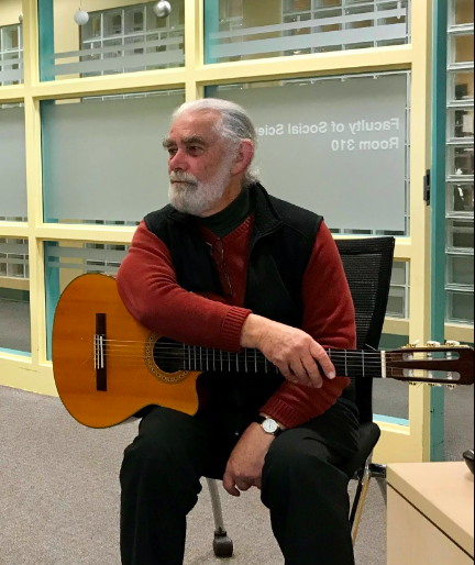 Dr. Tony Robertson, an older man with grey hair and beard, sits with his guitar.
