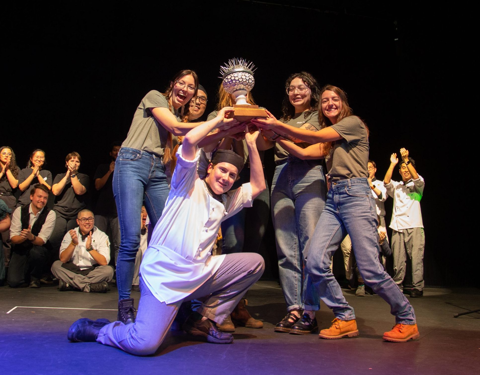 Six students holding a gray urchin-shaped trophy on stage with more students clapping behind them