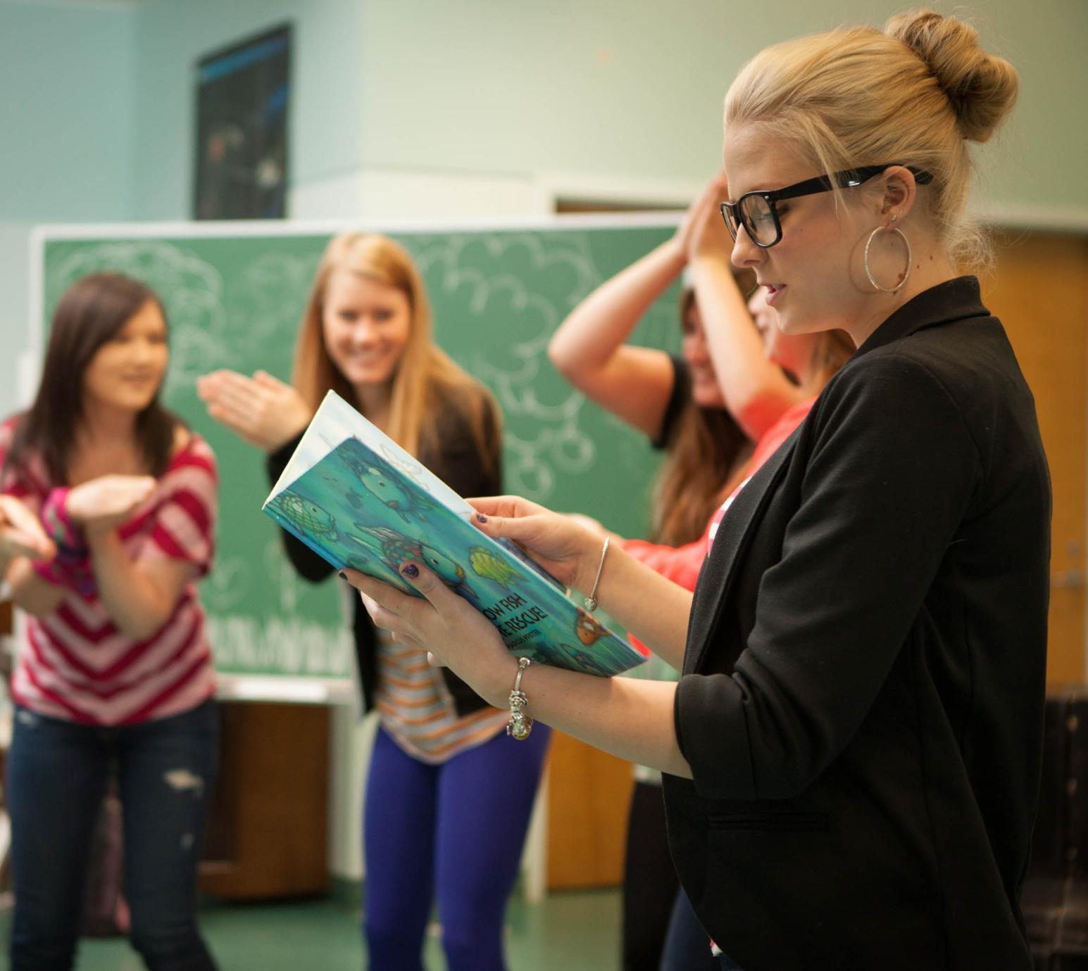 A person in glasses reads from a children's book as students listen behind her